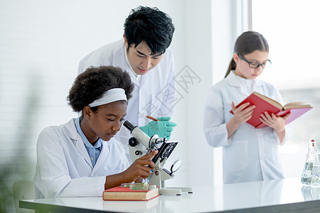 亚洲教师帮助并指导非裔美国年轻科学家女童使用显微镜分析实验室或教室中的植物组织 而其他学生则阅读一些书籍 校对 Portnoy/图片