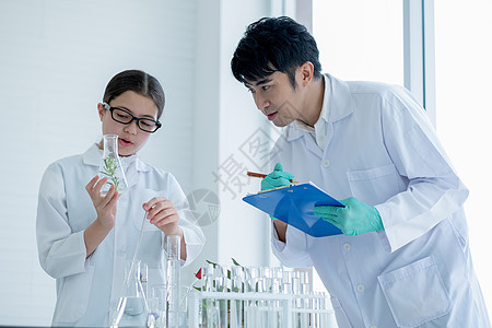 年轻的白人科学家女童拿着装有植物组织的烧瓶 并在实验室或教室做实验时向她的亚洲老师解释并记录一些数据图片