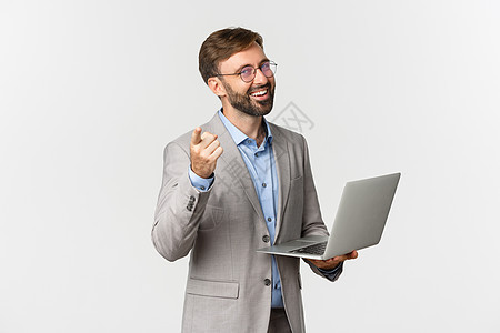 穿着灰色西装和眼镜的快乐而愉快的商务人士肖像 与笔记本电脑合作并用手指对着相机 赞美好的想法 站在白色背景之上经理职业套装老板促图片