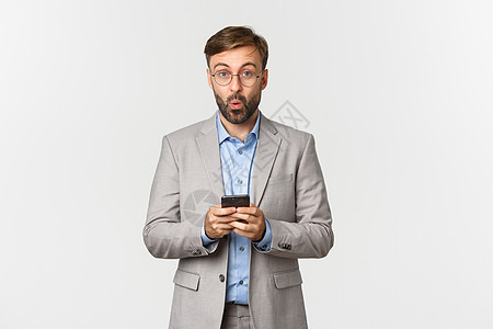 留着胡子 穿着灰色西装和眼镜的英俊商务人士的肖像 在手机上读了一些有趣的东西后看起来很有趣 站在白色背景上人士微笑套装工作管理人图片
