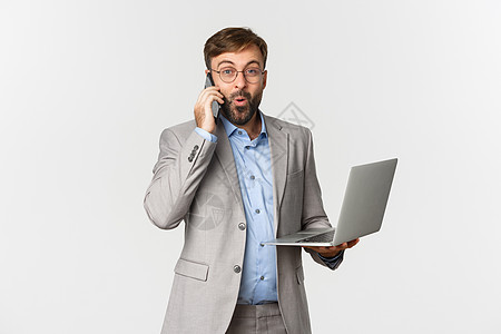 穿着灰色西装和眼镜的英俊商务人士肖像 在打电话时收到好消息 持有笔记本电脑 看着满意 站在白色背景上商业工作金融商务企业家人士快图片