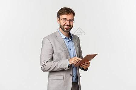 穿着灰色西装和眼镜的英俊胡子男性企业家肖像 使用数字平板电脑 工作 在照相机上微笑快乐 站在白色背景之上情感管理人员套装广告职业图片
