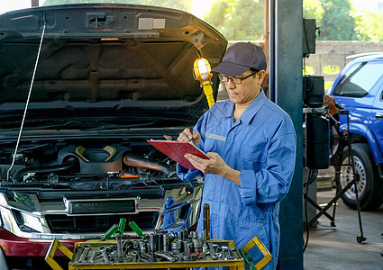 亚洲汽车机械师的肖像和蓝色制服 检查机头名单 解决车库中的车辆问题 30图片