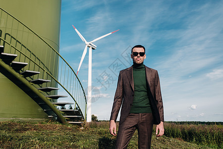 在田野和蓝天的背景下 一个身穿西装 身穿绿色高尔夫衬衫的男子站在风车旁边 风车附近的商务人士 未来的现代概念发电机银行家天空高尔图片