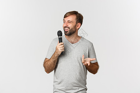 身穿灰色T恤衫的有魅力男性喜喜喜剧演员制作演讲 握着麦克风和说话 站在白色背景上广告商业衬衫胡子快乐唱歌胡须发型标识微笑背景