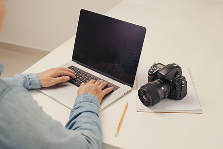 年轻男子摄影师在计算机上工作 手持键盘 相机 笔记本电脑和镜头的工作台设计师办公室职业单反工作室编辑桌子照片摄影爱好图片