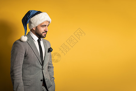 穿着蓝色圣诞老人帽子和西装的时髦英俊帅哥 看着在黄色背景中被孤立的相机 积极情绪 感情季节性节日派对男人套装乐趣商务人士新年季节图片