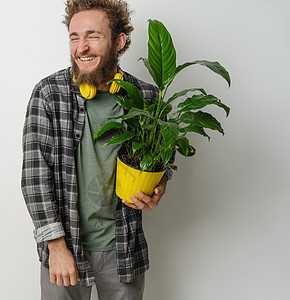 英俊微笑的留着胡须的年轻男子拿着黄色花盆 脖子上挂着穿着格子衬衫和黄色耳机的植物 与白色背景隔离 移动概念生物男性成人零售植物学图片