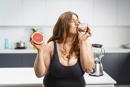 年轻的胖女人拿着蛋糕和葡萄柚选择吃什么 美丽胖乎乎的年轻女人吃不健康的食物 吃蛋糕和吃水果的胖女孩比较图片