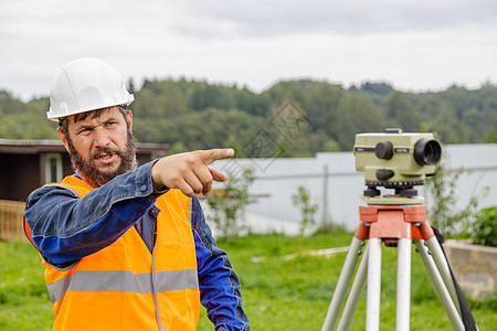 一位具有光学水平的土木工程师 用他的手从远处控制着同事绿色场地地面建设者男性施工电平工具望远镜检查图片