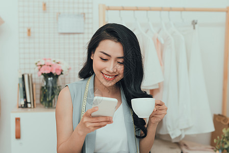 亚洲年轻女商务人士时装设计师在喝咖啡和使用手机时休息的画像桌子电话女性生意作坊纺织品办公室工作室衣服人体图片