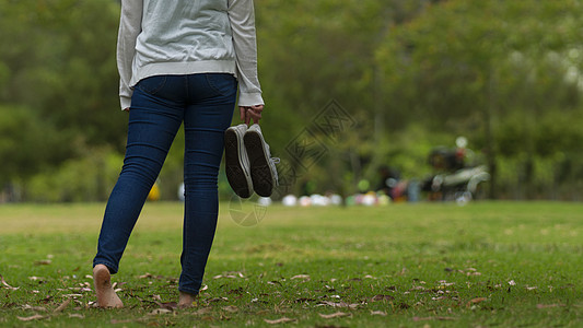 在公园赤脚裸足的青年女子近视中 她们用绿树背景将运动鞋握在右手上图片