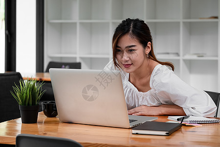 微笑的亚洲女学生使用笔记本电脑与老师在线学习 快乐的年轻女性学习语言听讲座看网络研讨会写笔记看笔记本电脑坐在咖啡馆里 远程教育技图片