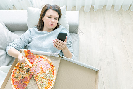可怜的女人吃披萨 拿着电话 在家里沙发上躺着 蓝调焦虑手机心碎女性房子蓝色悲伤女孩互联网细胞背景图片