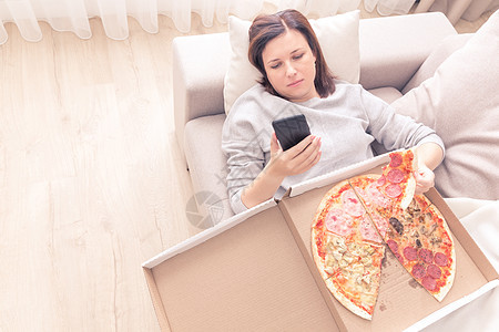 压抑的女人吃披萨 拿着电话 在沙发上铺在家中 温暖的黄色音调图片