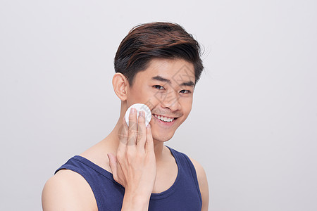 英俊的男子清洁脸皮皮肤 在灰色背景上用棉垫击打棉花和照相机图片