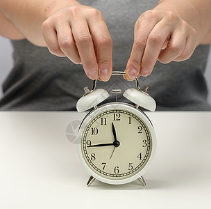 女性手在白桌子上握着一个白色金属闹钟 时间是15至12时 任务期限的构想 分配的期限图片