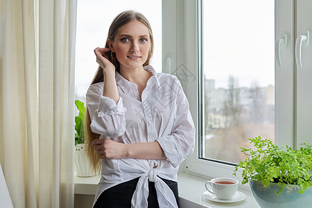 在冬季秋天窗口附近 看着摄像机的金发年轻女人的肖像幸福房子公寓成人金发女郎窗户微笑乐趣快乐闲暇图片