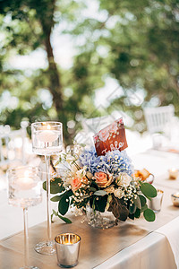 贵宾接待中心婚礼餐桌招待会 花瓶中的花束放在桌子上 上面放着粉色和白色的玫瑰和蓝色绣球花 盘子上有桌子的编号 在高玻璃烛台上燃烧蜡烛背景