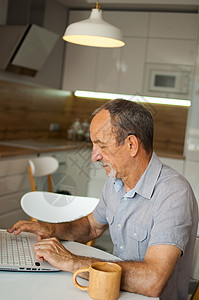 Trendy成熟男子在家工作 手持笔记本电脑坐在厨房的桌子上 享受退休快乐 家庭办公室概念图片