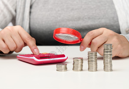 一个女人坐在桌子旁 用计算器计算 另一只手拿着放大镜 桌子上有一叠硬币 费用和收入的计算图片