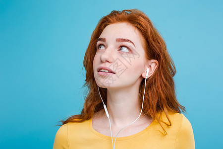 生活方式概念 — 欢快快乐的姜红头发女孩的肖像喜欢戴着耳机听音乐 对着镜头欢快地微笑 孤立在蓝色柔和的背景上 复制空间图片