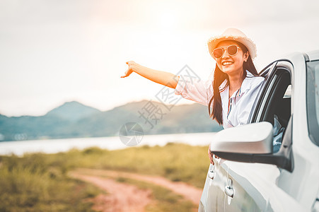 快乐的女人在草地和高山湖泊背景的开窗车外挥手 人们在假期的公路旅行中放松作为旅行者的生活方式 运输和旅行概念手臂天空日落驾驶阳光背景图片