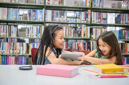 两个女孩一边看书一边在教室里争夺平板电脑 人们的生活方式和教育 学校概念中的青年友谊和儿童关系 日托和多样性主题幸福苗圃朋友姐姐图片