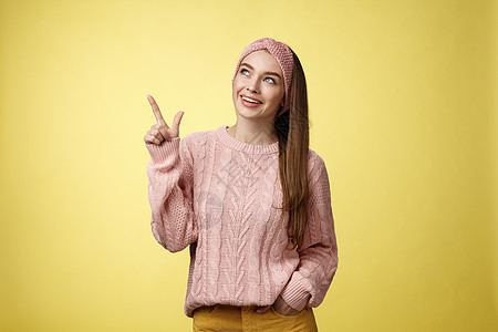 有趣有趣的可爱 20 多岁欧洲女孩的肖像穿着毛衣 头带看着左上角指向侧面微笑着好奇 欣喜若狂 被黄色墙壁上的好奇促销捕获顾客情感图片