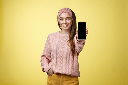 最好的智能手机在这里 迷人外向的年轻漂亮女性穿着针织毛衣 头带伸出手 手机显示小工具屏幕 在黄墙上微笑着推荐手机软件大学福利细胞图片