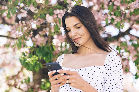 在樱花树的背景下 在公园里使用智能手机的美丽女人的画像 使用社交网络浏览互联网的年轻美女店铺阳光樱花销售量网页动作细胞工作服务考图片
