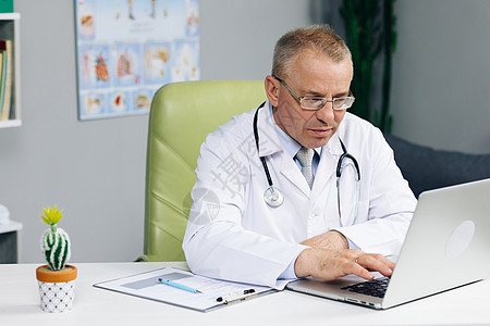穿着白色实验室大衣的医生正在医院办公室的桌子后面浏览病史 戴眼镜的冷静家庭医生正在健康诊所的笔记本电脑上工作图片