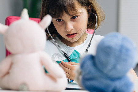 可爱可爱的学龄前小女孩穿着医疗制服拿着听诊器听生病的玩具病人 聪明的小孩玩医院游戏作为医生假装护士的概念图片