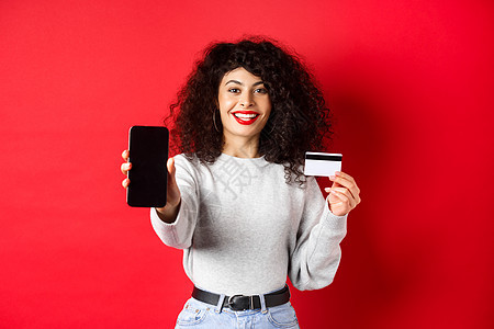 展示塑料信用卡和空智能手机屏幕的快乐漂亮女孩 展示账户或应用程序 站在红色背景上广告购物技术卷曲工作室黑发生活女性情感电话图片