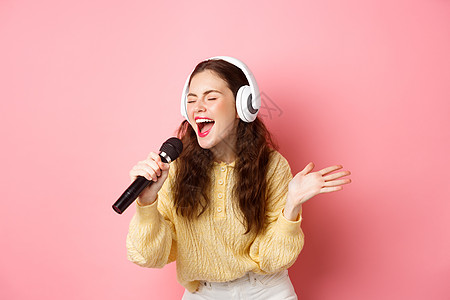 年轻女性唱卡拉OK 戴耳机和表演歌曲 握着麦克风 站在粉红背景下站立的肖像快乐唱歌手势女孩工作室女士卡拉ok魅力歌手购物图片