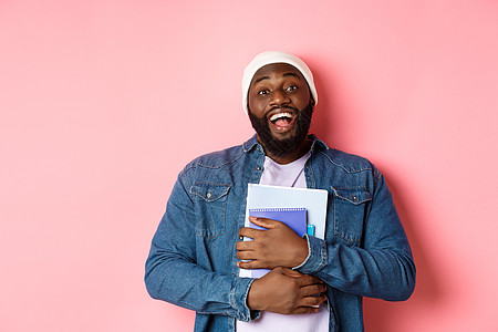 教育 非洲美国美籍大豆男学生快乐 拥有笔记本 学习课程 对着镜头微笑 站在粉红背景上潮人帽子成人工作室衬衫培训班男人购物横幅广告图片