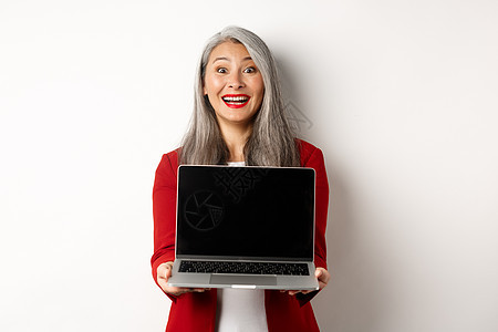 快乐的高级女企业家展示了空白笔记本电脑屏幕 对着镜头微笑地微笑 站在白色背景上站立雇主商业经理头发成功人士成人化妆品嘴唇外套图片