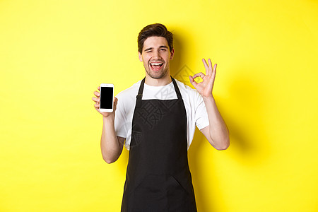 咖啡店的英俊工人在黄色背景上站立 展示OK牌子和智能手机屏幕 推荐申请以及服务员微笑咖啡师男性电话快乐广告互联网推销员成功图片