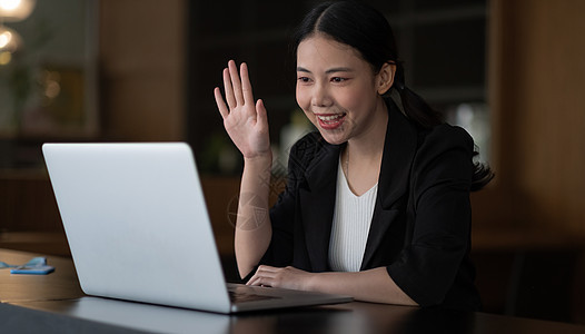 身着白衬衫的亚裔女性在办公室用视频通话或聊天时 在笔记本电脑上微笑和挥手图片