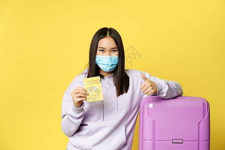 戴着面罩微笑的亚洲女游客 带着手提箱站着 为旅行者出示 covid 国际疫苗接种证书 并竖起大拇指 黄色背景保健护照乘客肺炎假期图片