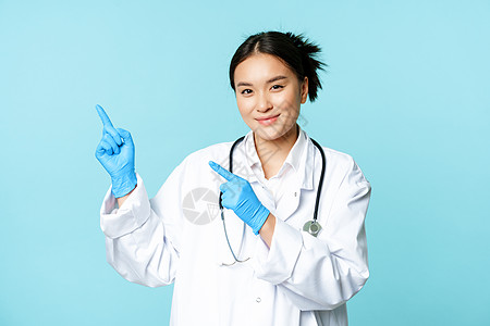 微笑的女医生 身穿医疗制服和无菌手套的医生 指着促销时留下的手指 诊所标志 站在蓝色背景上感染症状女性肺炎社交女士预防装备医师广图片