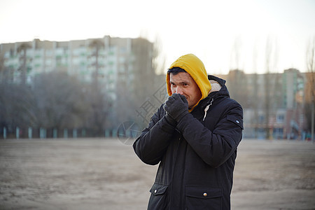 身穿黄帽和黑夹克的年轻冷冻男子 成年男性在寒冷的天气中用手套亲手取暖背景街道黑色呼吸男人空气闲暇拳头休闲服装图片