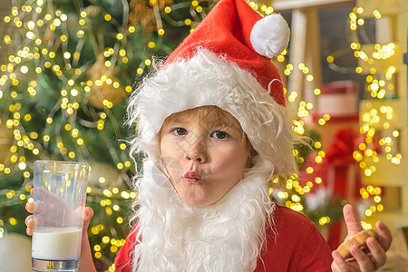 圣诞老人在圣诞树背景上拿着饼干和一杯牛奶 圣诞老人拿着圣诞饼干和牛奶反对圣诞树背景 小男孩圣诞老人图片