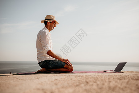 一个数字游牧者 一个戴帽子的男人 一个带着笔记本电脑的商务人士在日落时分在海边的岩石上做瑜伽 从远处在线开展业务 假期远程工作图片