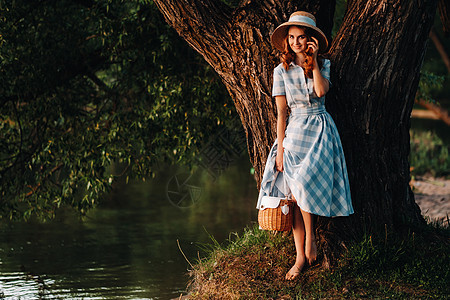 一个年轻美丽的女孩 戴着草帽和稻草袋 享受着湖的景色帽子头发农村日落草地男人公园紫色篮子花园图片