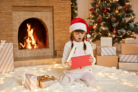 小女孩在圣诞节打开礼物时兴奋不已 女孩戴着圣诞老人的帽子坐在地板上 张着嘴 惊讶和惊讶 在客厅里摆出圣诞装饰的姿势图片