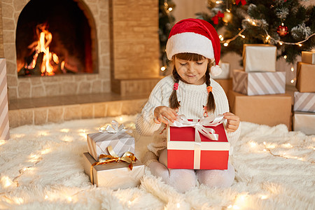 小女孩打开圣诞礼物 手指放在丝带上 微笑着看着礼品盒 孩子戴着圣诞老人帽在靠近壁炉和圣诞树的地板上摆姿势图片