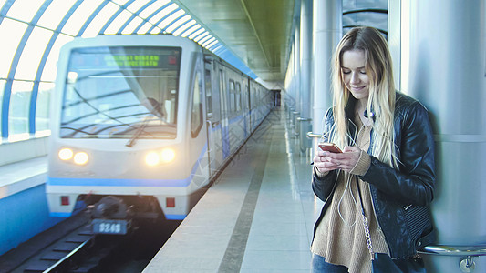 穿着皮夹克的金发长头发的女孩 在火车即将到来的背景下 站在地铁站直道上 从一个角度看图片