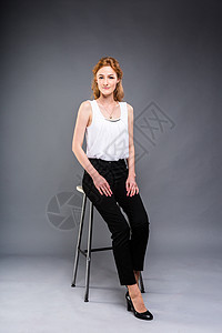 美丽的年轻白种女人 长着红色的长发 穿着高跟鞋 坐在椅子上 黑色的裤子和白色的衬衫在灰色的背景下完全成长 经营理念商业金发女郎肤图片