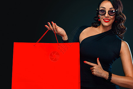 黑周五商店的销售概念 戴墨镜的购物女孩拿着红包在黑暗背景中被孤立 女性指着看左侧复制空间女士黑色快乐头发黑发购物奢华购物者购物中图片